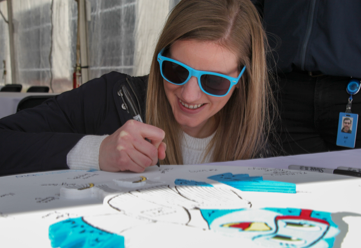 Girl wearing blue sunglasses bent down at a table writing with a marker