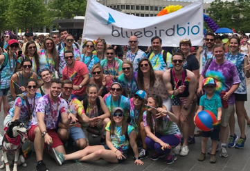 A group of people all wearing tie dyed t-shirts posed on the side of a parade route with a bluebird bio banner